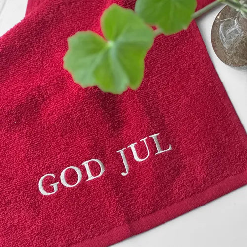 Julröd gästhandduk med texten God Jul broderad i vitt placerad på en badrumsbänk.
