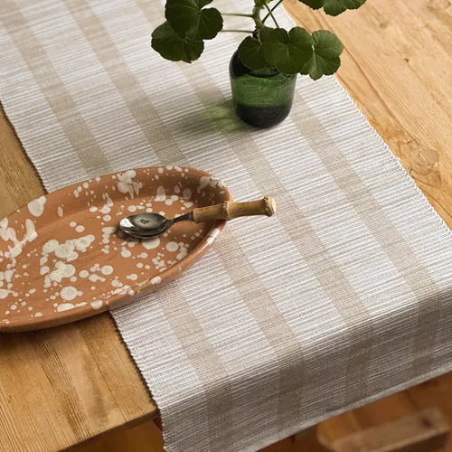 Randig bordslöpare i vitt och linnefärgat av återvunnen textil på träbord med keramikfat och krukväxt