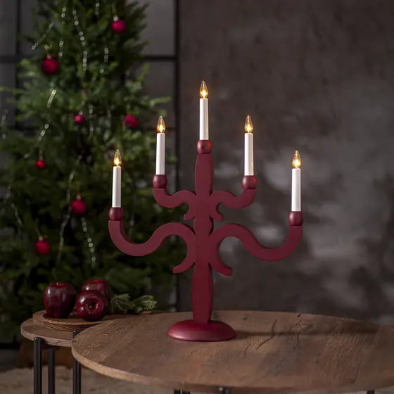 Djupröd adventsljusstake Canna med fem tända ljus på ett träbord framför en julgran med röda julkulor.