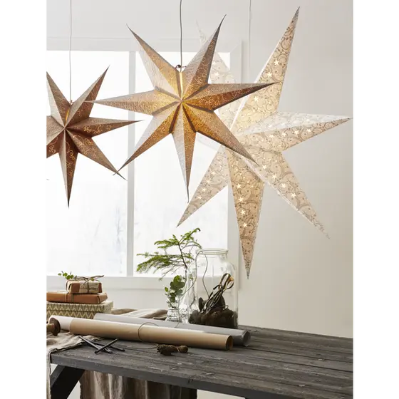 Guldig adventsstjärna Decorus med dekorativt hålmönster upphängd i ett fönster med varmt ljussken tillsammans med flera andra stjärnor.