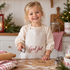 Ett barn i treårsåldern bakar pepparkakor iklädd ett linnefärgat barnförkläde med röd broderad text "God Jul" och ficka