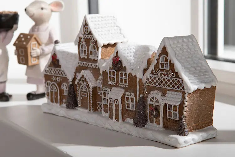 LED-lykta med fyra sammanbyggda pepparkakshus, snötäckta tak och vita dekorativa detaljer, placerad på en fönsterbräda brevid andra juldekorationer.