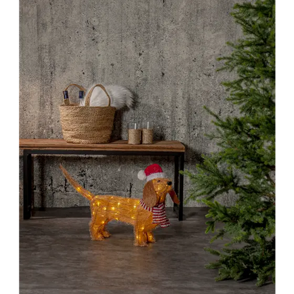 Lysande julhund med tomteluva och rödvit halsduk placerad intill en julgran i en stämningsfull julmiljö.