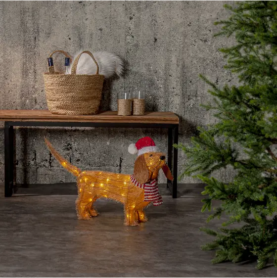 Lysande julhund med tomteluva och rödvit halsduk placerad intill en julgran i en stämningsfull julmiljö.