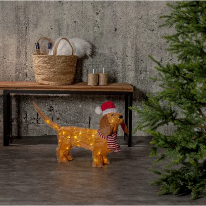 Lysande julhund med tomteluva och rödvit halsduk placerad intill en julgran i en stämningsfull julmiljö.