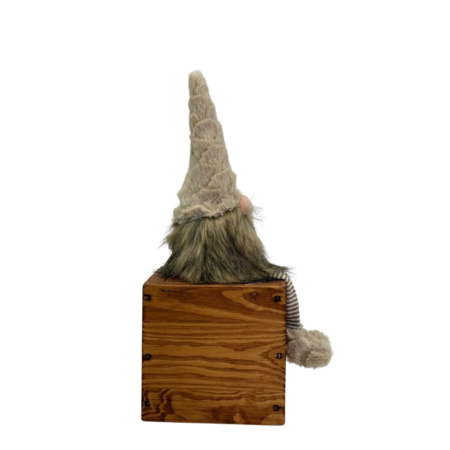 Liten tomte i naturfärger som sitter och dinglar med benen över en hyllkant från Miljögården, sett från sidan