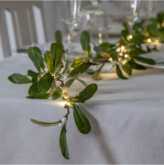Närbild av mistelgirlang med gröna blad och varmvita LED-ljus längs kanten på ett dukat bord med vit duk.